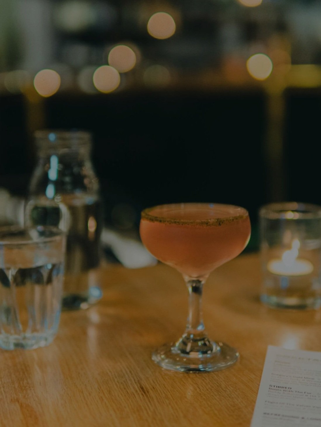 A dimly lit orange cocktail in a coupe glass sits on a table while lights from the bar are out of focus in the background